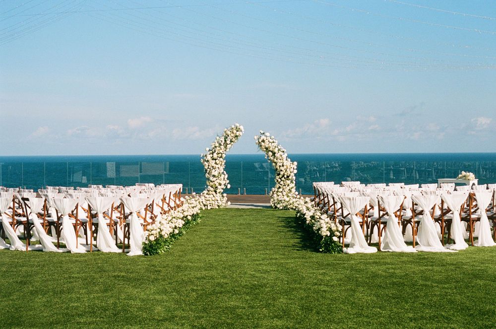 Wedding ceremony set up outdoors with white chairs arranged in rows overlooking the ocean on a bright sunny day.