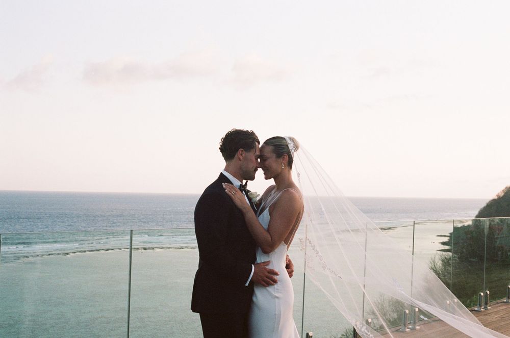 Romantic couple embrace on a coastal cliff during sunset with ocean waves crashing in the background.