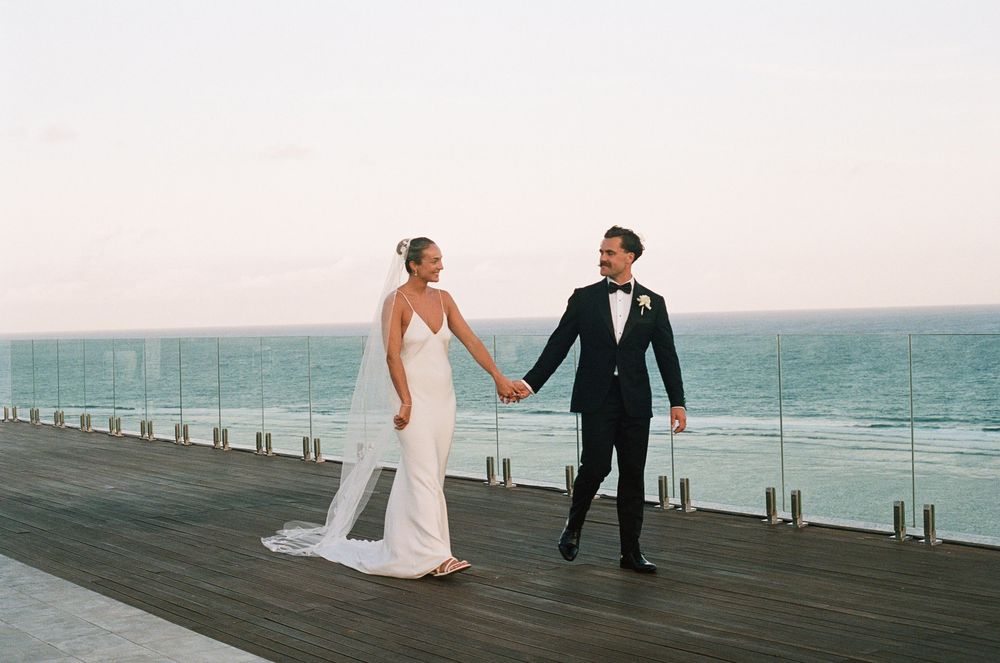 A couple in wedding attire holding hands while walking along a seaside boardwalk during sunset.