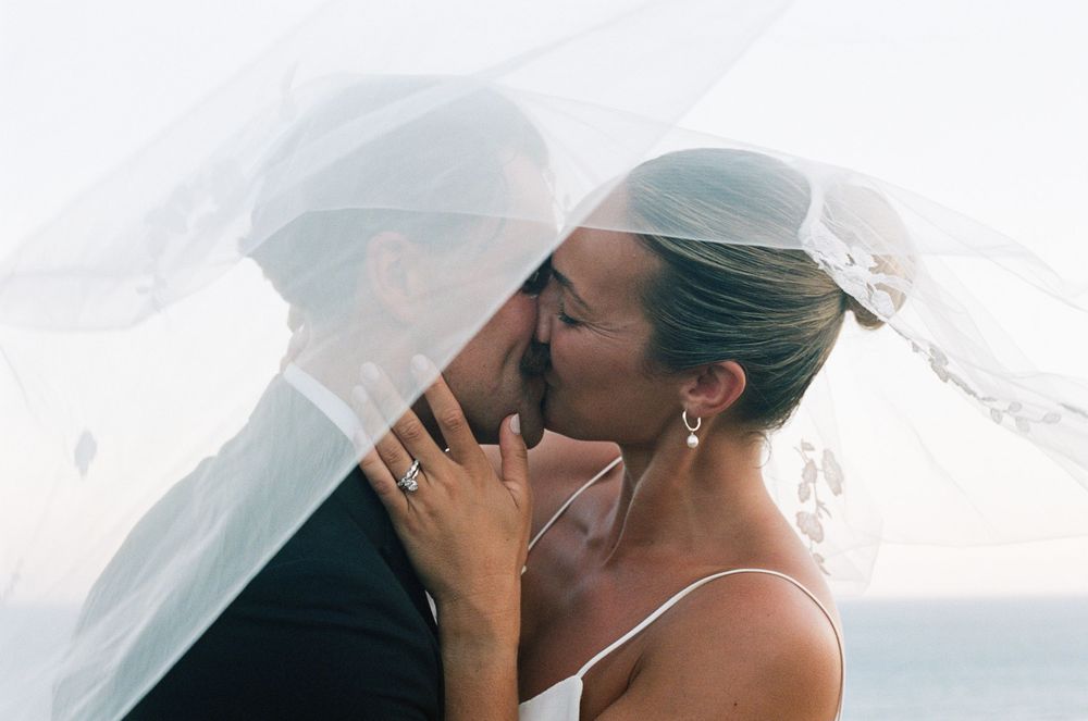 A tender moment captured under a flowing veil as a bride and groom share a kiss on their wedding day.