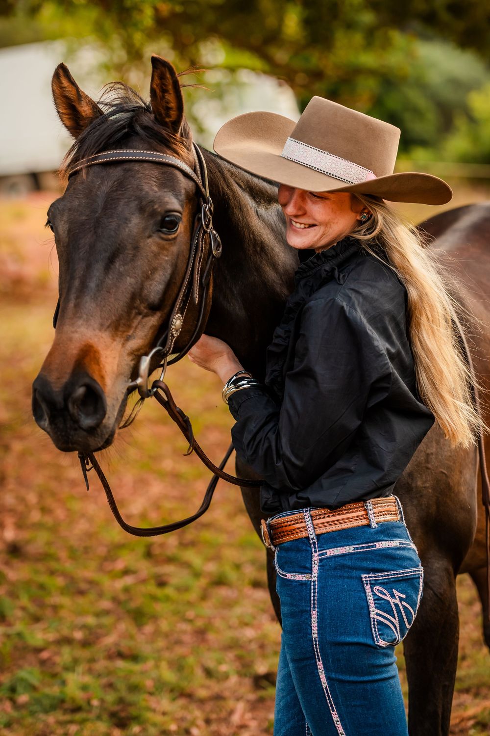Dark horse with black mane stands beside rider wearing blue jeans and brown cowboy hat in autumn setting.