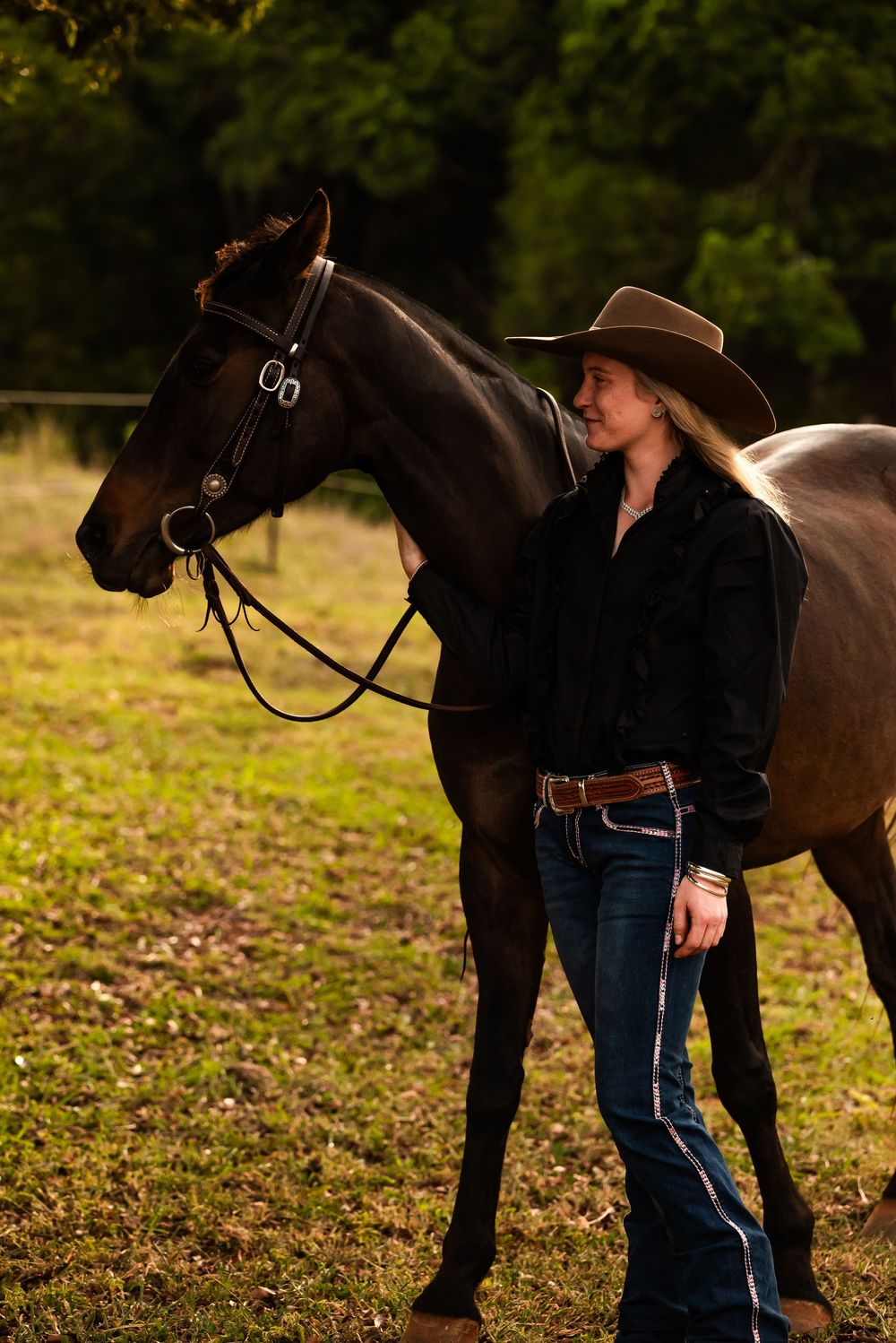 Person in blue jeans tending to a dark brown horse in an autumn field during afternoon light.