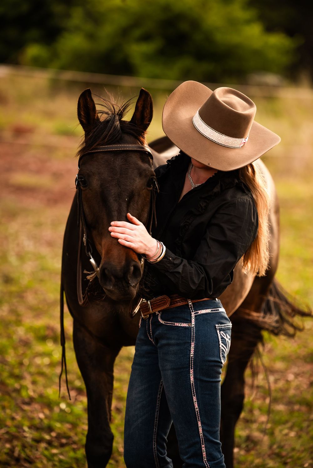 A person wearing a brown cowboy hat and black jacket stands next to a dark horse in an outdoor setting.