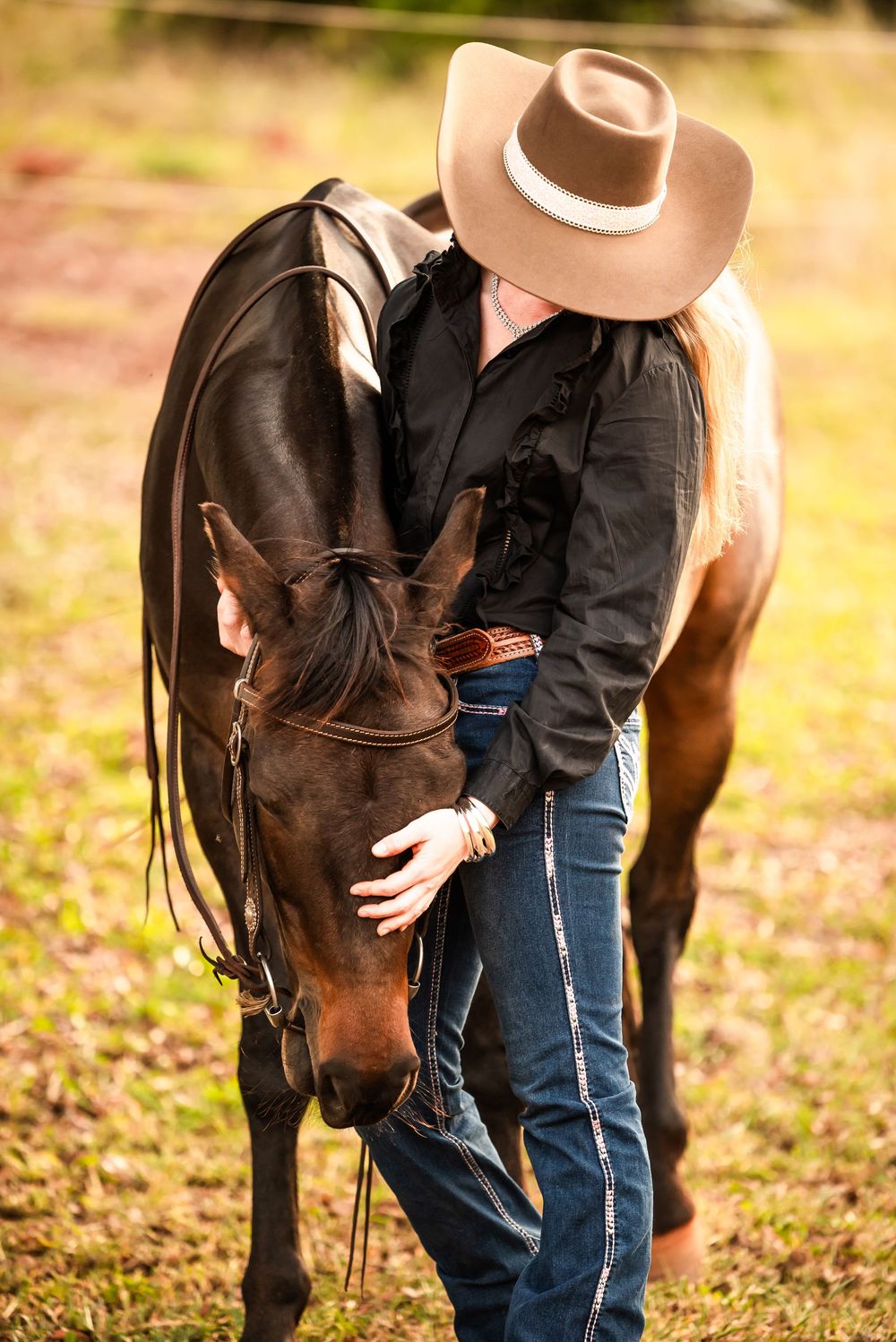 Person in cowboy hat and jeans stands beside dark brown horse in fall outdoor setting during golden hour.