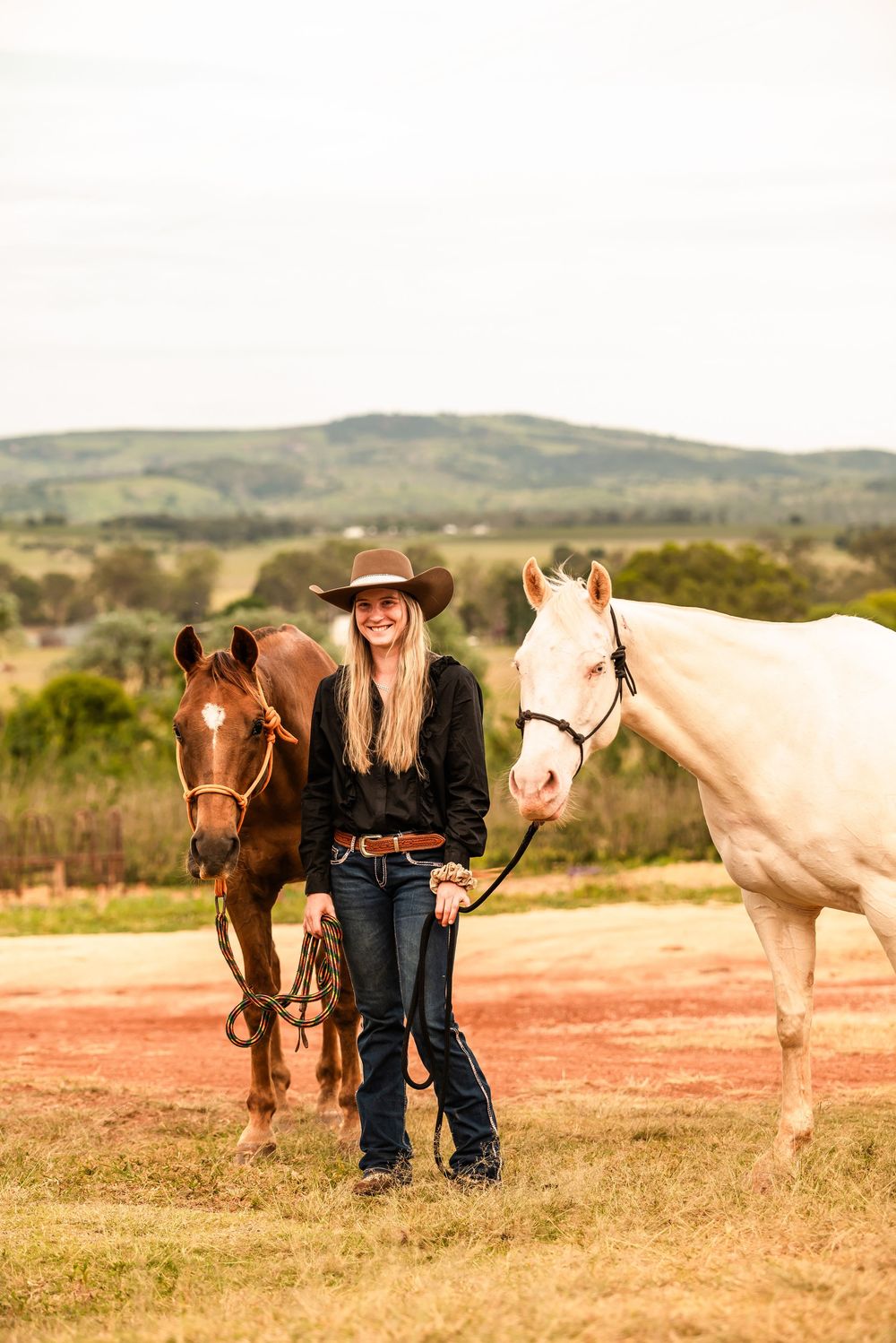 White and brown horses graze in a pastoral field while someone in western attire stands with them at sunset.