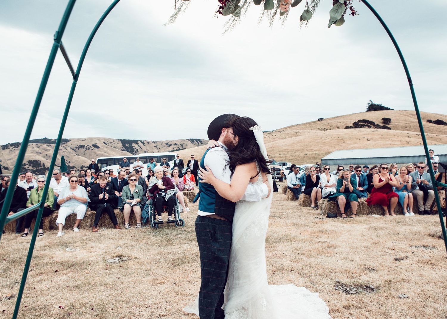 A couple shares a romantic moment during their outdoor wedding ceremony under an arch with guests watching in the background.