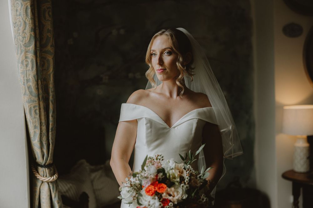 A bride in a white dress holds a colorful bouquet while getting ready in a sunlit room.