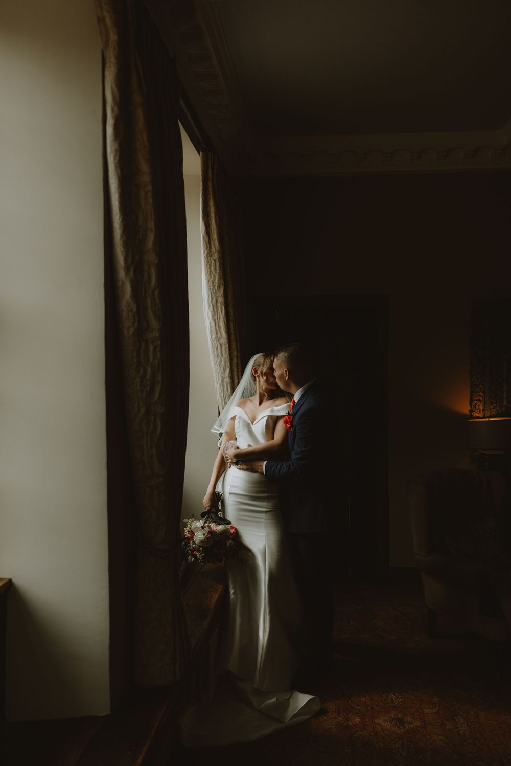 Moody artistic shots of a bride standing by a window in dramatic lighting.