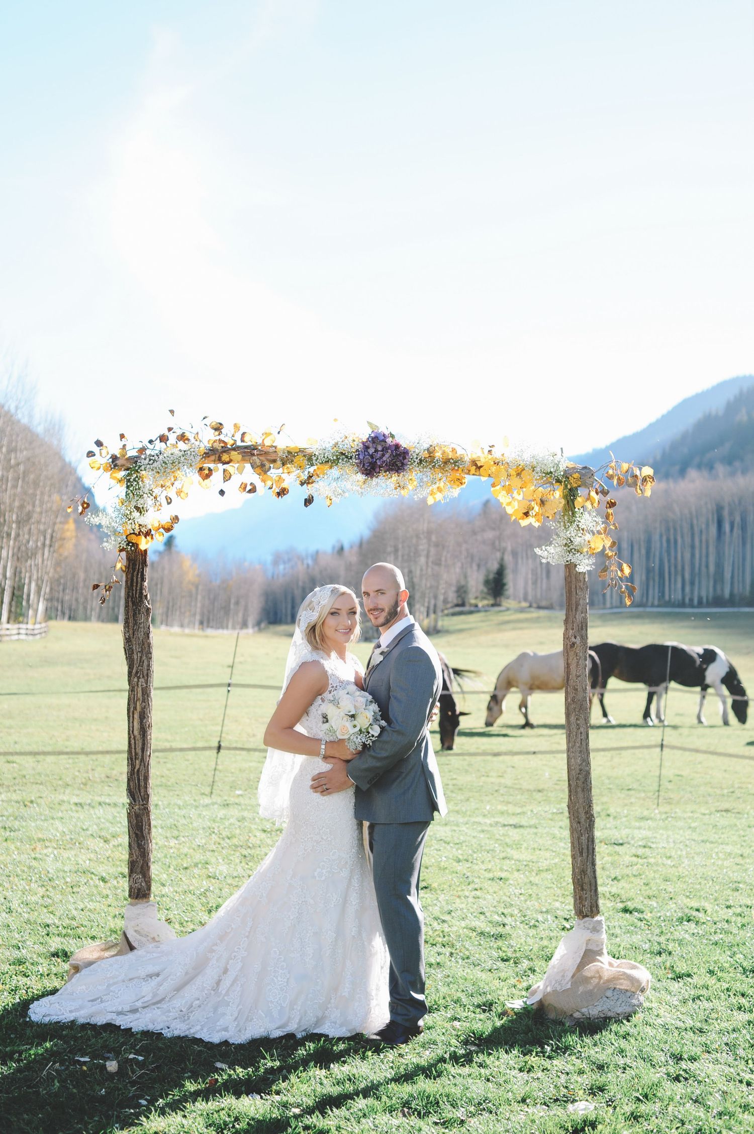 Wedding couple poses under floral arches in a mountain meadow with horses in background.
