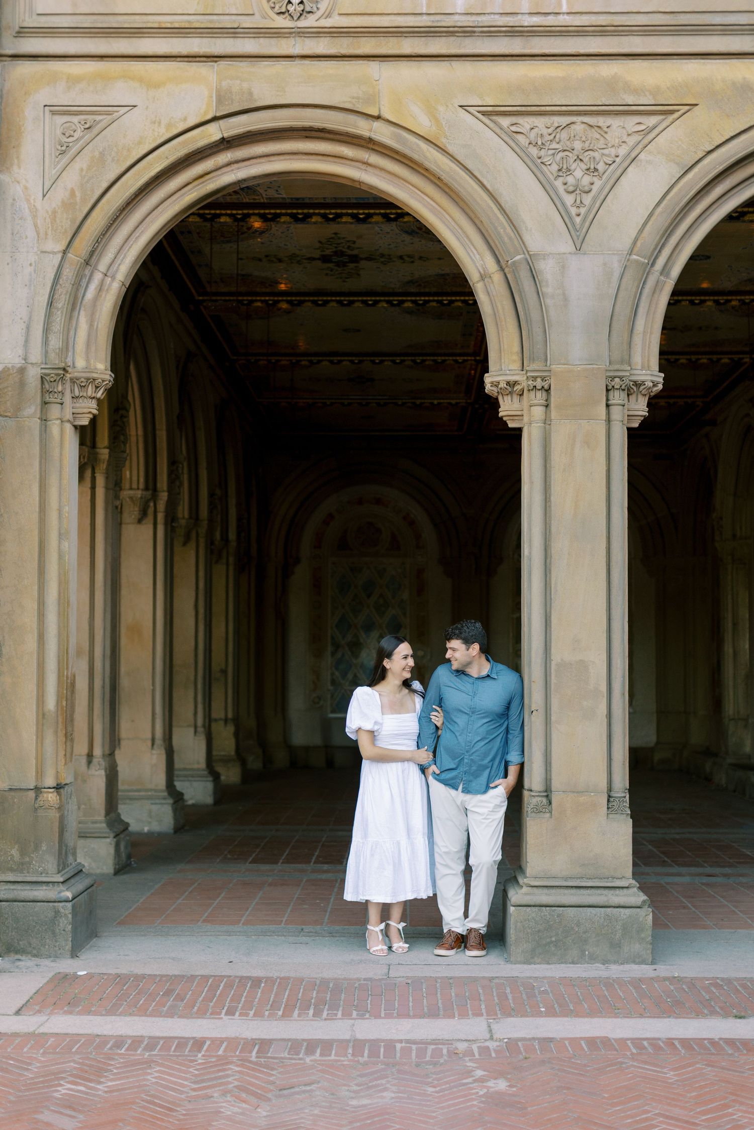 A couple stands in a graceful archway of a historic building with ornate stone architecture and brick flooring.