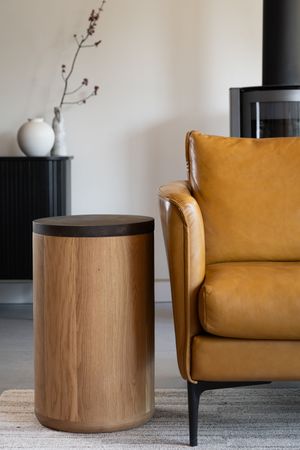Stylish round wooden side table with dark top displayed next to a tan leather armchair on gray carpeting with minimalist decor.