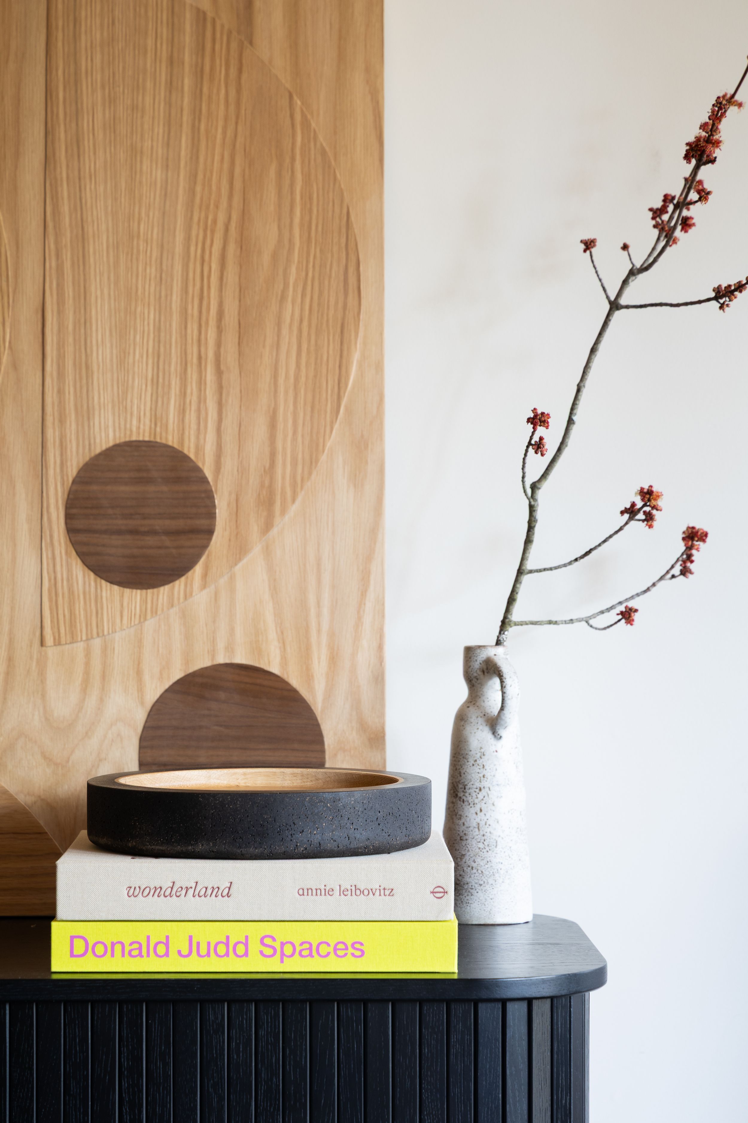 Minimalist home decor with wooden art pieces, design books, and a vase with delicate flowering branches on black cabinet.