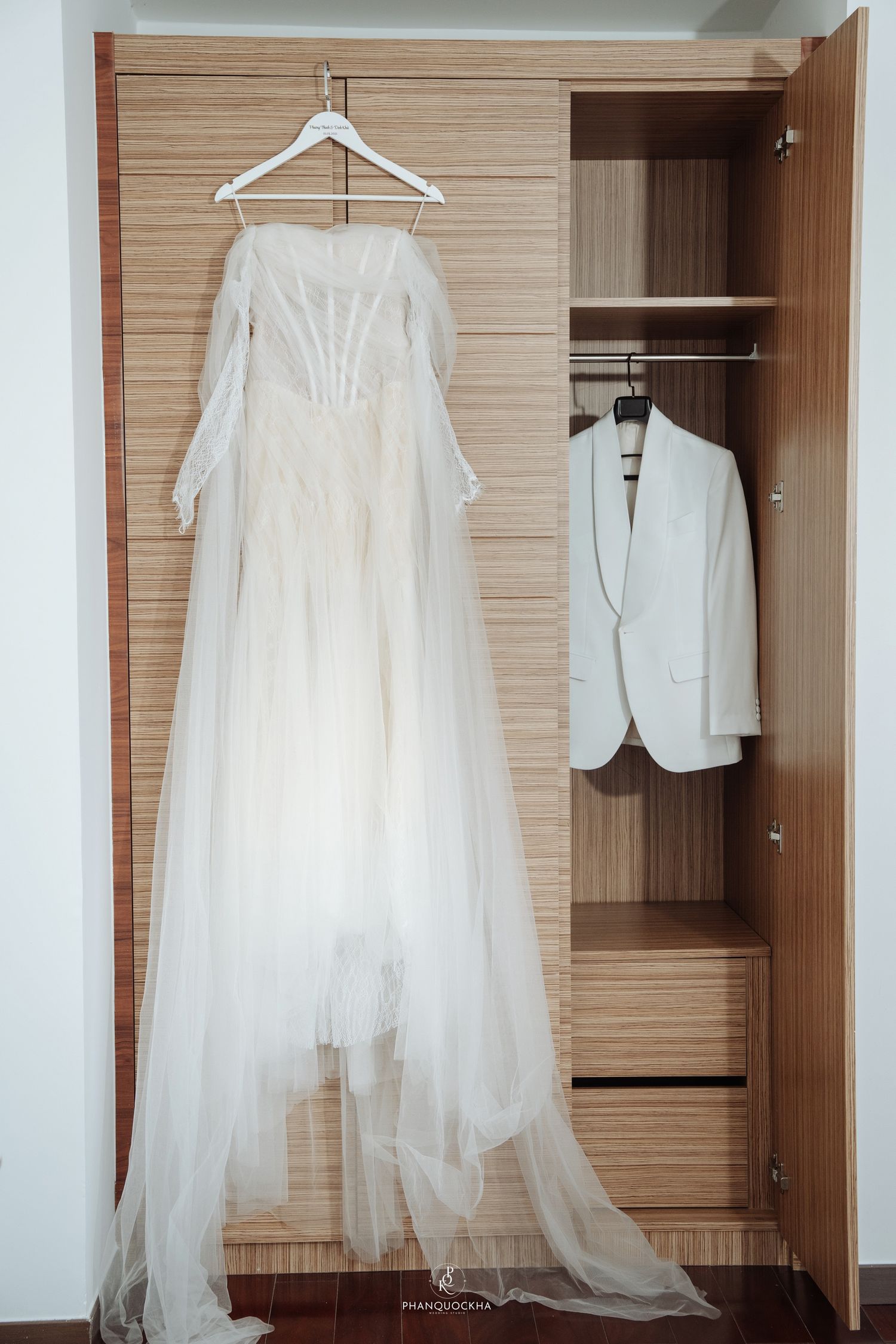Wedding dress and white groom suit jacket hanging side by side in a wooden wardrobe closet.