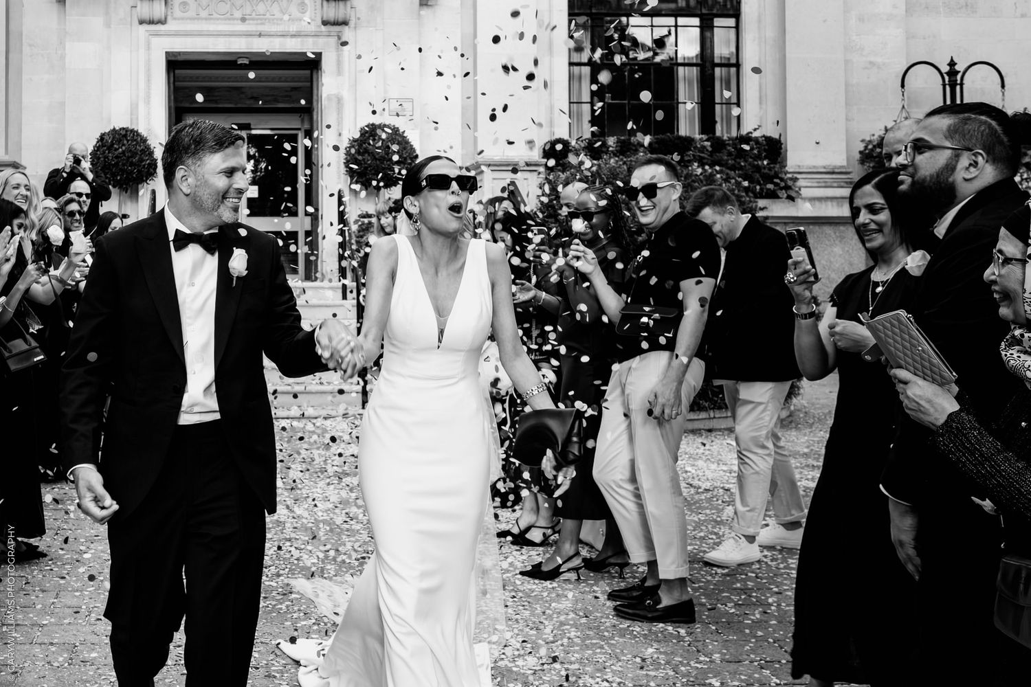 Wedding couple exits venue while guests throw confetti in black and white photograph captured at outdoor celebration.