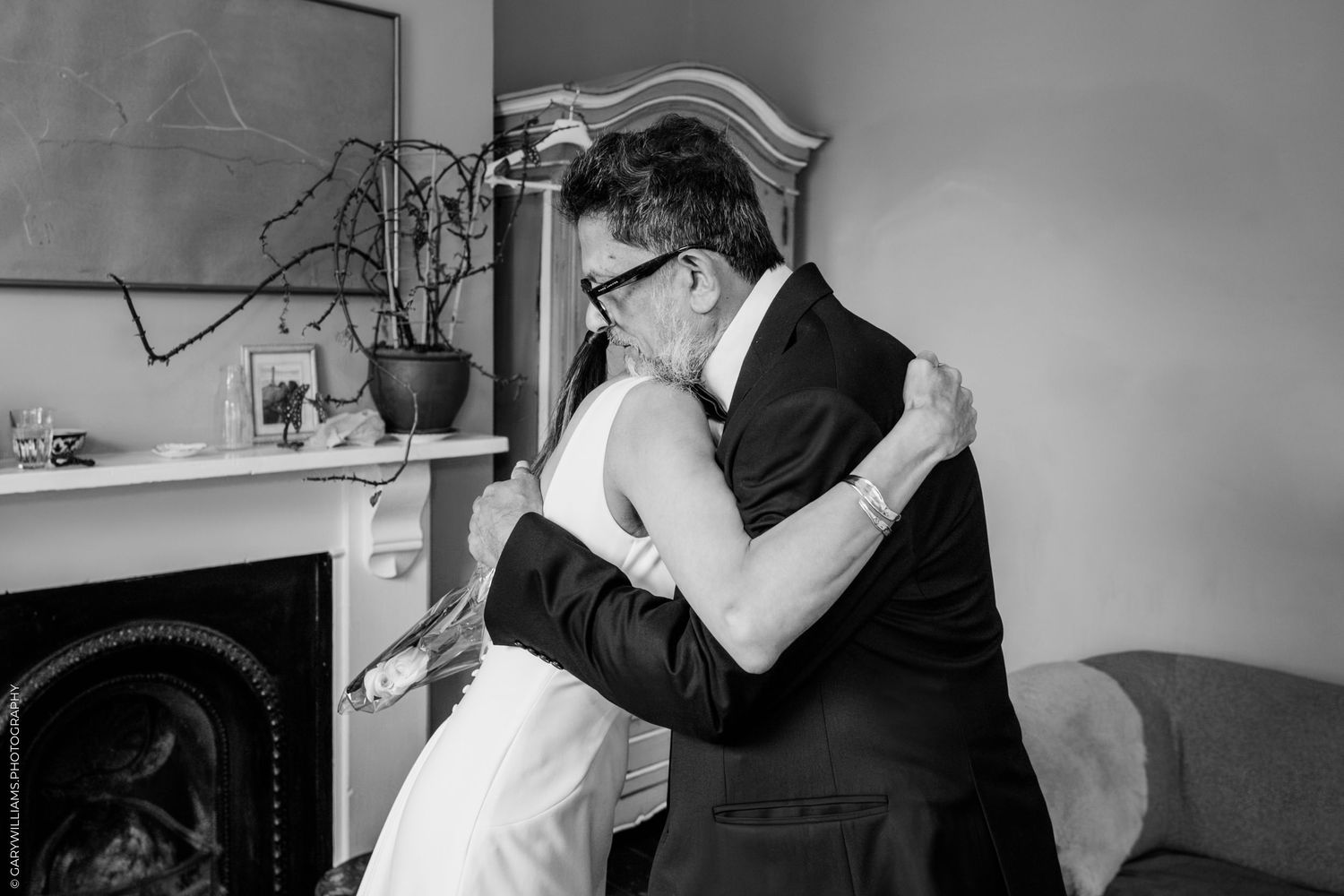 A tender embrace between two people at a wedding, captured in elegant black and white photography next to a fireplace.
