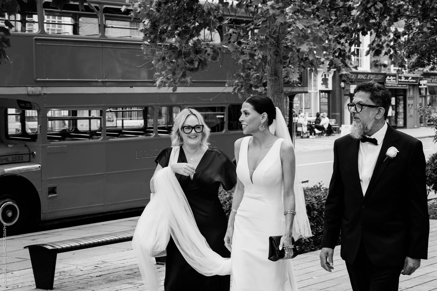 A joyful black and white wedding photograph showing the bridal party walking on a London street with a red bus in background.