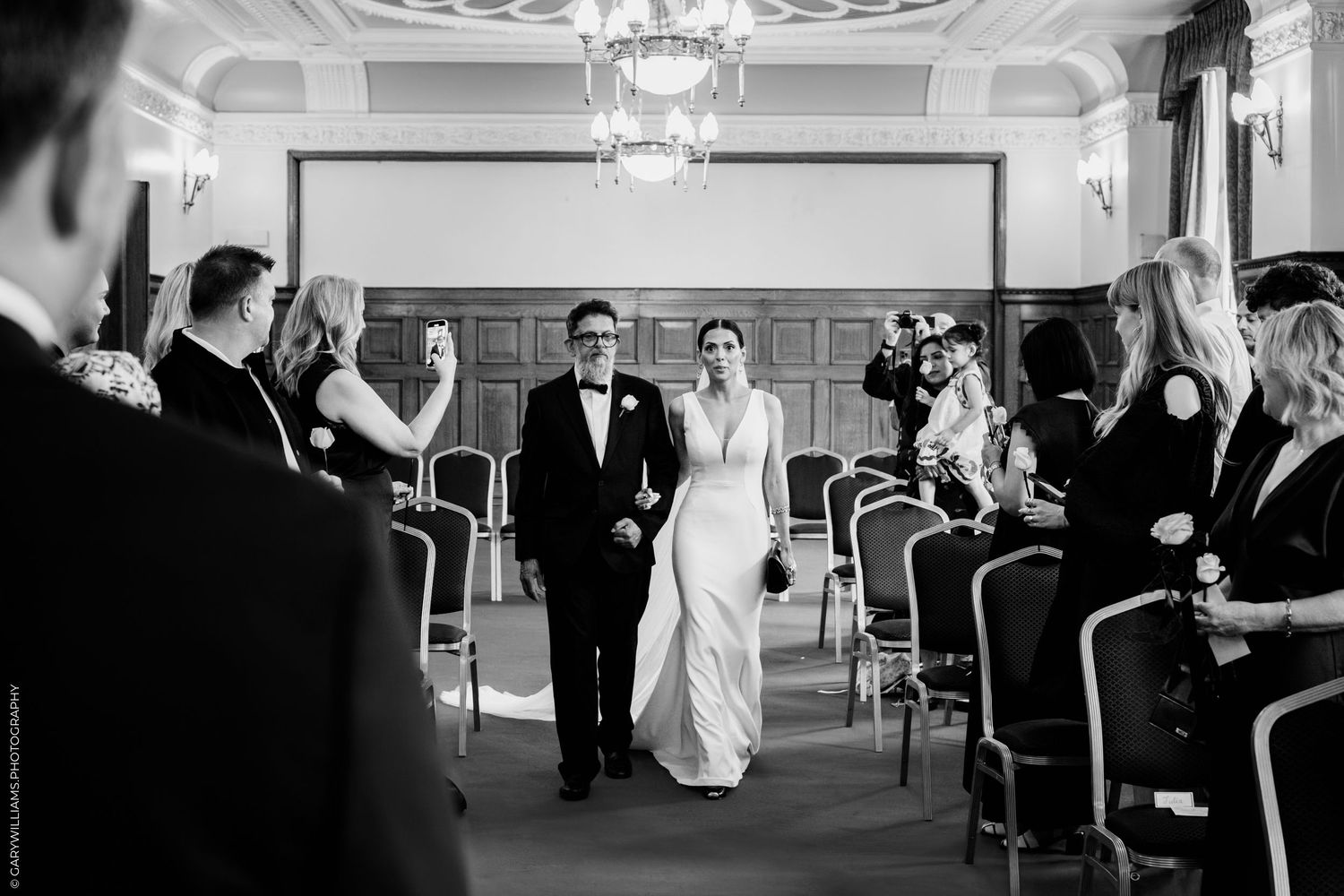 Wedding ceremony procession in a grand ballroom with chandelier and guests lining both sides of the aisle.