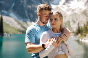 Couple in casual attire embrace while enjoying scenic views of a mountain lake on a sunny summer day.