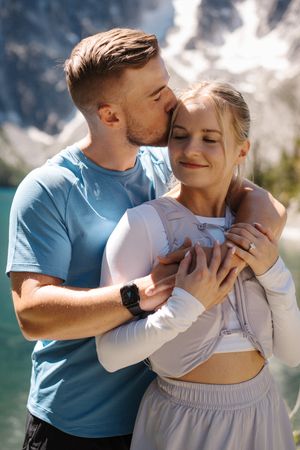 A couple embraces lovingly against a scenic mountain backdrop on a cloudy day wearing blue and white clothing.