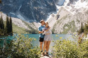 Couple standing at edge of Lake Moraine with turquoise waters and snow-capped Rocky Mountains in background.