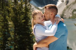 A tender embrace between two people against a scenic mountain lake backdrop with evergreen trees in the sunlight. Colchuck Lake proposal