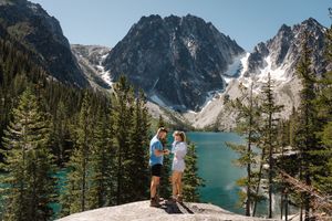 Two people enjoying the view of turquoise Colchuck Lake surrounded by snowy peaks and evergreen trees in Washington State.