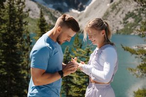A couple in casual attire share romantic moments during a lakeside photoshoot at a scenic mountain location.