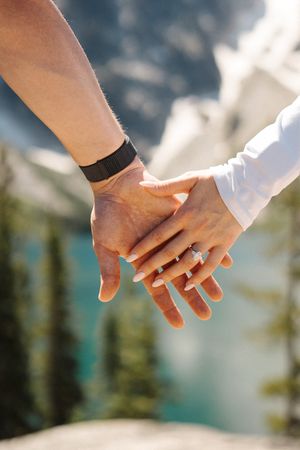 Two hands reach out to hold each other against a blurred mountain and forest landscape background. Colchuck Lake proposal