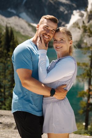 Couple embracing by a scenic mountain lake with turquoise water and rocky cliffs in the background. Colchuck Lake proposal