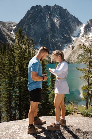 A couple looks at their phones while standing on a rocky outcrop overlooking a stunning mountain lake and peaks.