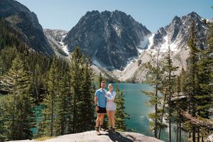 Two figures stand on a rocky overlook admiring a turquoise alpine lake surrounded by snow-capped mountains and evergreens.