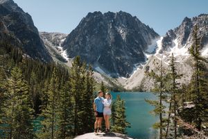 Two people embrace while standing on a rocky overlook above turquoise Colchuck Lake surrounded by snow-capped peaks.