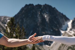 Two hands reaching toward each other against a backdrop of a majestic snow-capped mountain peak under blue skies.