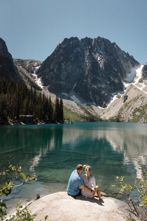 Two people sit together on a rock overlooking a turquoise alpine lake surrounded by snow-capped mountains Colchuck Lake proposal