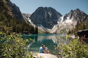 Snow-capped mountain peaks tower over a pristine alpine lake surrounded by evergreen forest on a sunny day. Colchuck Lake proposal