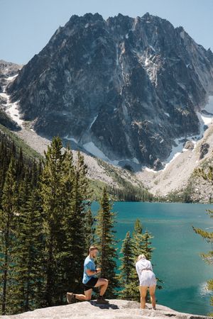 Two figures standing on rocky overlook above pristine alpine lake with dramatic mountain backdrop.