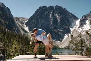 A couple performs a romantic dip pose against a backdrop of snow-capped mountains and alpine lake in the wilderness. Colchuck Lake proposal