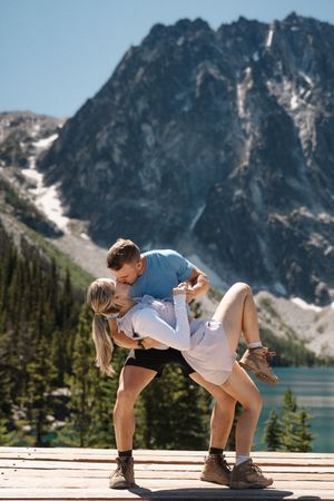 Couple shares a romantic kiss on a wooden deck overlooking a serene mountain lake and snow-capped peaks. Colchuck Lake proposal