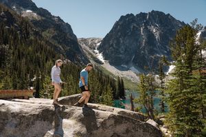 Two hikers stand on a rocky overlook with a dramatic mountain lake and snow-capped peaks in the background. Colchuck Lake proposal