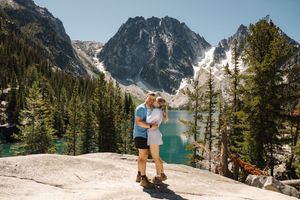 A couple embraces while standing on a rocky overlook above Colchuck Lake proposal with snow-capped mountains in Washington State.