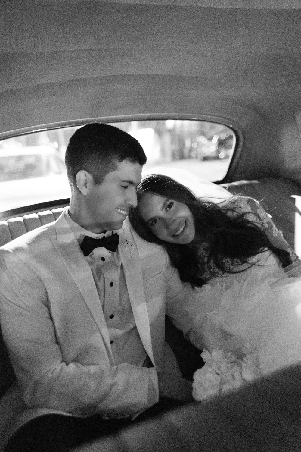 A romantic black and white wedding photo of newlyweds sharing a tender moment together in the backseat of a car.