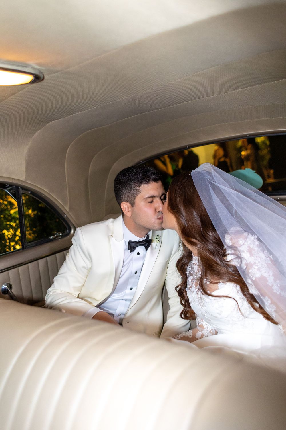 A newlywed couple shares a romantic moment in the back of a luxury vintage car with cream leather interior.