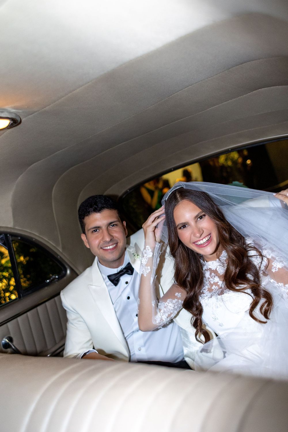 A newly married couple sits together in the backseat of a classic car, with the bride wearing a lace gown and veil.