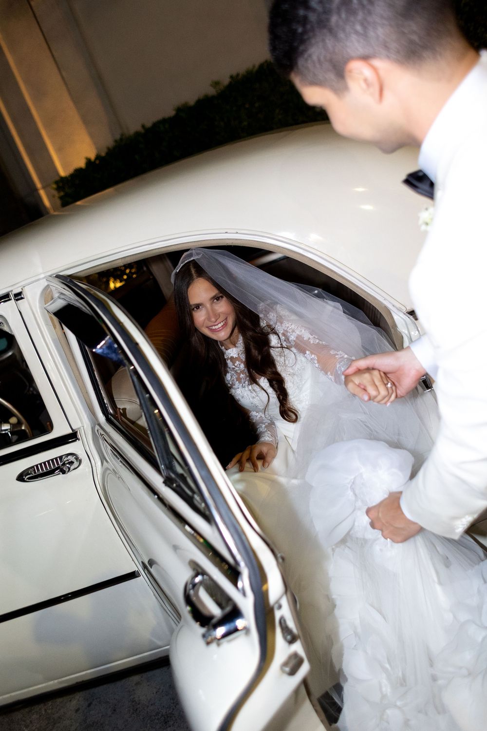 A bride in a white wedding dress being helped into a white car during a wedding exit sequence.