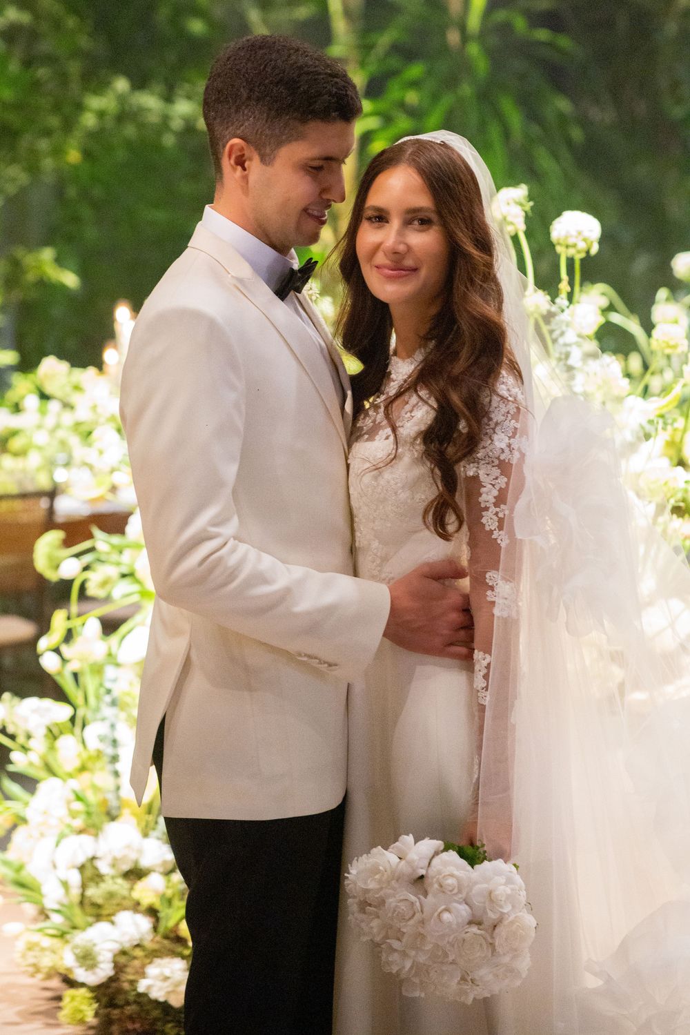 A bride and groom share an intimate moment in their white wedding attire surrounded by flowers in a garden setting.
