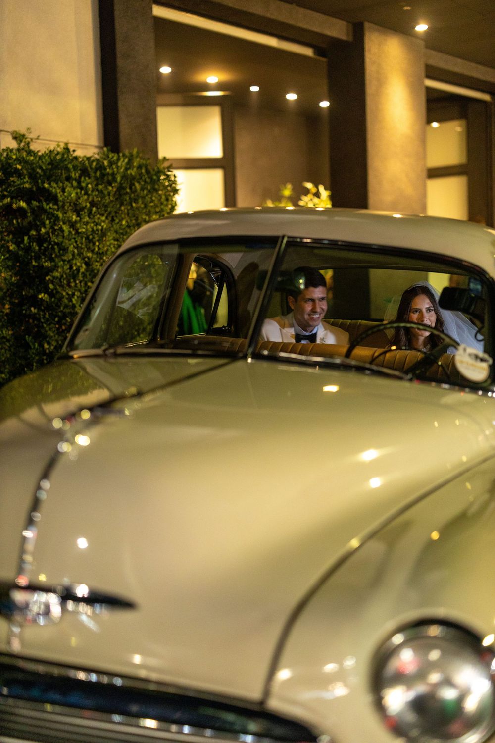 Four sequential views of a classic cream-colored vintage luxury car parked under lights in an evening setting.