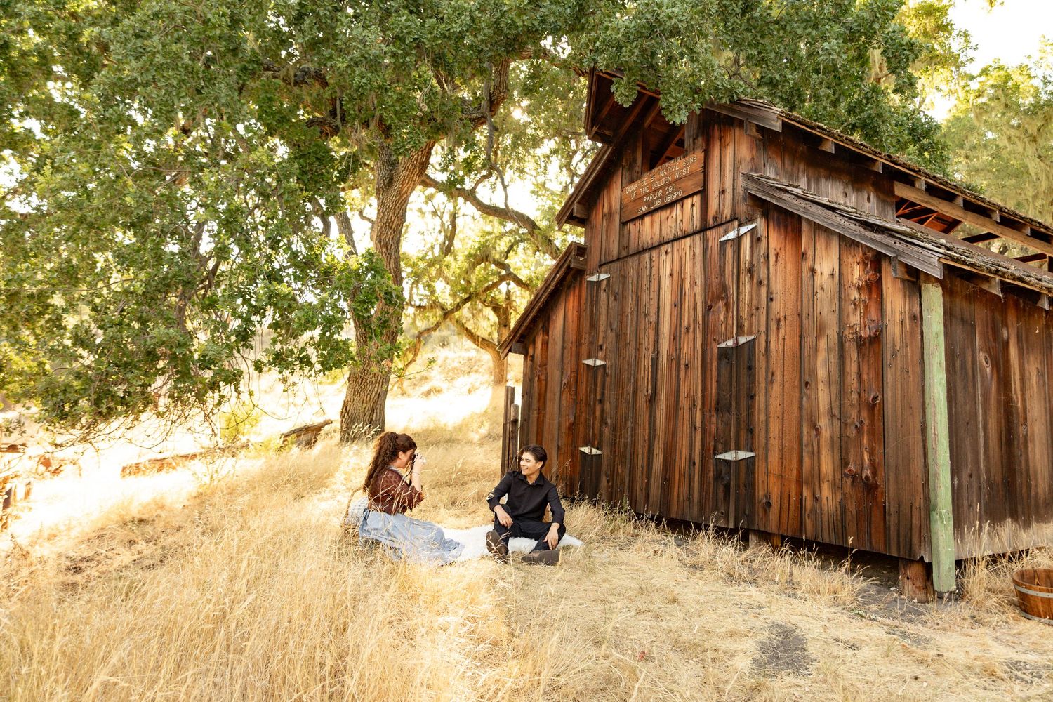 A rustic wooden barn sits among golden grass and oak trees during sunset in a rural California landscape.