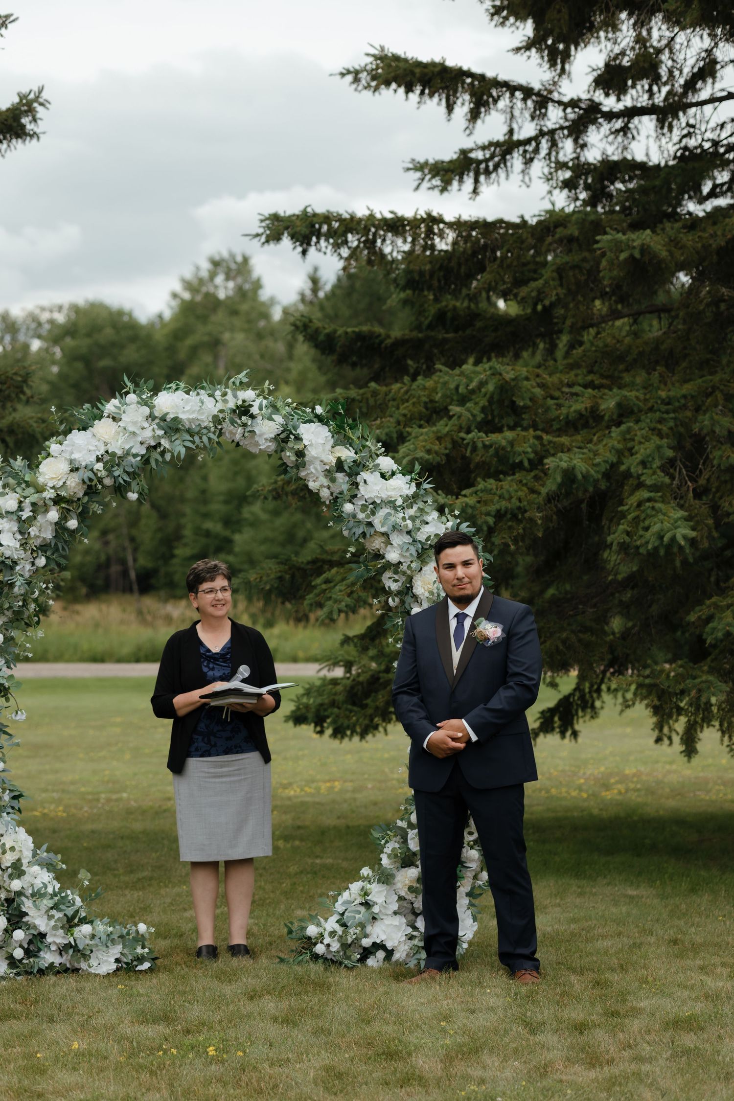 Wedding party members pose playfully behind evergreen trees in an outdoor setting with a white floral arch.