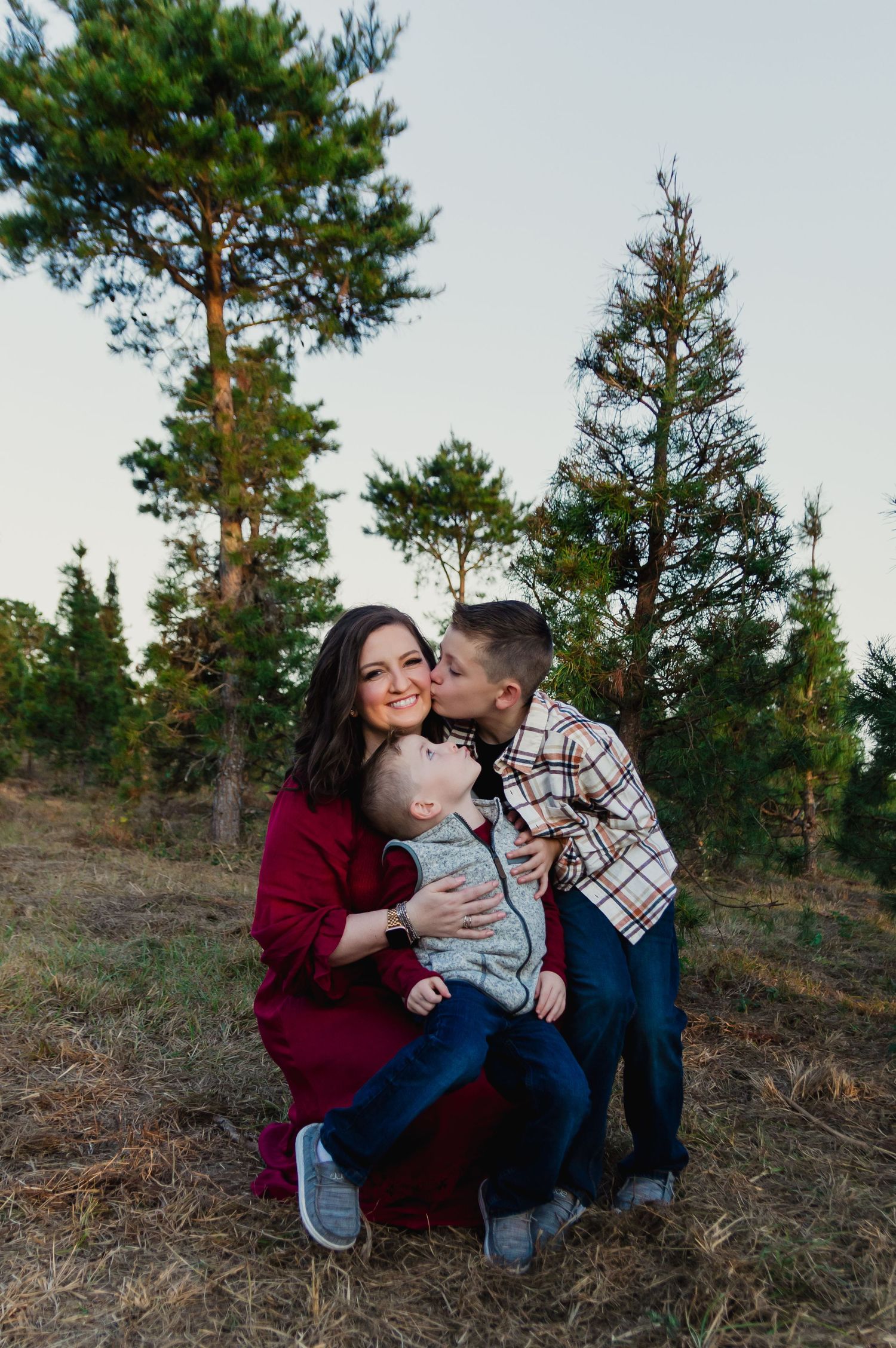 A family enjoying a tender moment together outdoors among tall pine trees against a light sky at dusk.