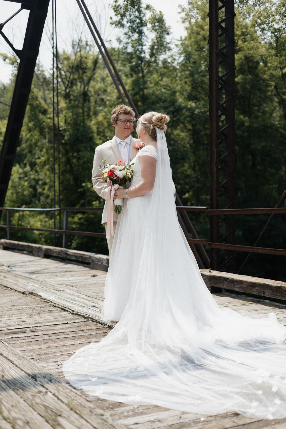 A wedding couple stands together on a rustic wooden bridge surrounded by green trees on a sunny day.